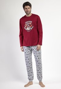 Hombre con una camiseta de manga larga roja con un batería de dibujos animados y pantalones de pijama grises con pequeñas caras de dibujos animados similares y patrones.