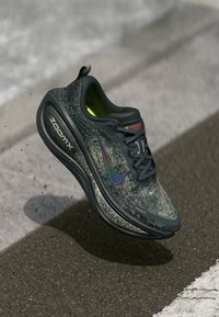 Dunkelgraue Nike ZoomX Laufschuhe mit schimmerndem Swoosh, schwarzen Schnürsenkeln und neon-gelbem Innenfutter in der Luft über einer Asphaltstraße.