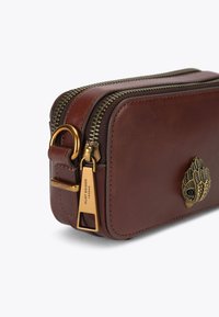 Sac en cuir marron avec des accents dorés, une fermeture éclair et un emblème décoratif en forme de main sur le devant. Texture lisse et forme rectangulaire.