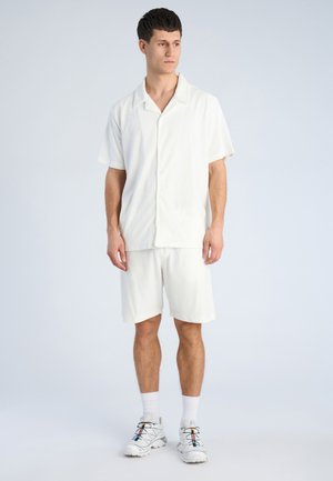 Ung mand står, iført hvid kortærmet knap-op skjorte, hvide shorts, hvide sokker og hvide sneakers med farverige snørebånd.