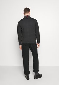 Maglione grigio scuro a maglia con colletto alto, caratterizzato da un accentuazione a righe rosse e bianche, abbinato a pantaloni neri e scarpe nere.
