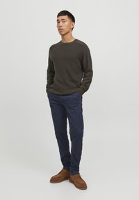 Dunkelolivestrickpullover mit langen Ärmeln, marineblaue Chinos und braune Wildlederschuhe. Das Modell steht mit den Händen in den Taschen vor einem hellen Hintergrund.
