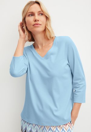 Blonde vrouw met lichtblauw V-hals shirt met driekwart mouwen en gebloemde broek, poseert met hand bij het oor, kijkt opzij.