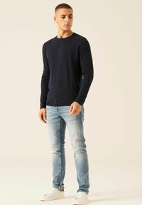 Marineblauer Langarmpullover mit strukturiertem Finish, kombiniert mit hellblauen Slim-Fit-Jeans und weißen Sneakers, in stehender Pose gezeigt.