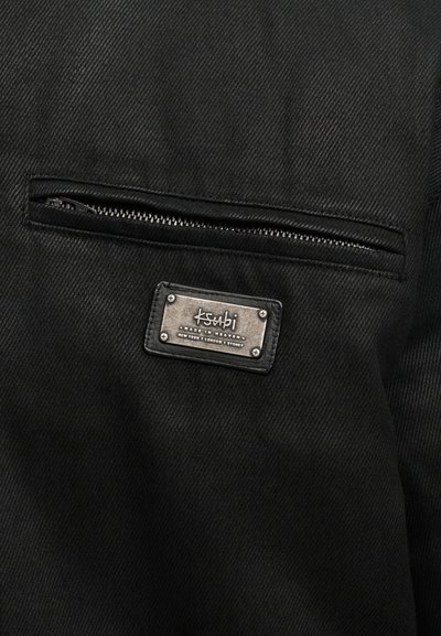 Μαύρο υφασμάτινο μπουφάν με τσέπη με φερμουάρ, υφή υφάσματος με ανάγλυφο πλέξιμο και μεταλλική ετικέτα μάρκας στερεωμένη με πριτσίνια.