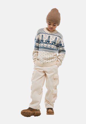 Enfant portant un bonnet beige en tricot, un pull crème avec un motif de phare et d'ancre bleu marine, un pantalon cargo crème et des bottes de randonnée marron, debout les mains dans les poches.