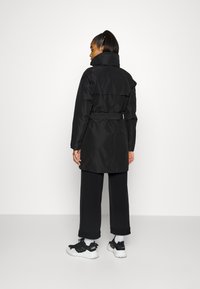 Czarny płaszcz z wysokim kołnierzem, ściąganym paskiem i długimi rękawami, wykonany z gładkiego materiału. Nosi się go na luźne czarne spodnie i sportowe buty.