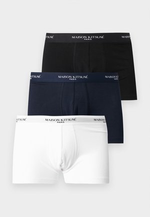 Młody mężczyzna o jasnej karnacji i brązowych włosach stoi boso, ubrany w czarne bokserki z logo „Maison Kitsuné” na białym tle.