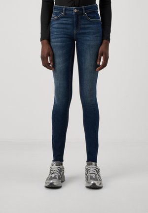 Vaqueros skinny en denim azul oscuro con una textura suave, cintura alta, cinco bolsillos y detalles de costura sutiles. Llevados con zapatos deportivos grises.