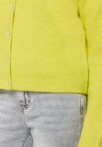 Żółty kardigan wykonany z miękkiego materiału, z przodu na guziki, z prążkowanym wykończeniem na dole i długimi rękawami. Zestawiony z szarymi jeansami w kropki.