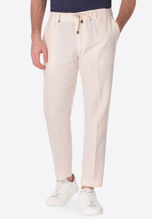 Uomo con pantaloni color crema con coulisse e sneakers bianche, mani in tasca, su uno sfondo chiaro e uniforme.