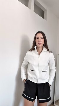 Fehér cipzáras dzsekit viselő nő, fekete szegélyű zsebekkel és fehér csíkos fekete sorttal, bent áll egy fehér fal előtt.