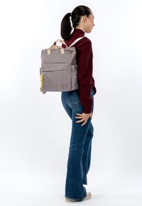 Szary plecak o teksturowanej powierzchni, z dwoma bocznymi kieszeniami, górnymi uchwytami i regulowanymi beżowymi paskami. Posiada żółty suwak i logo.