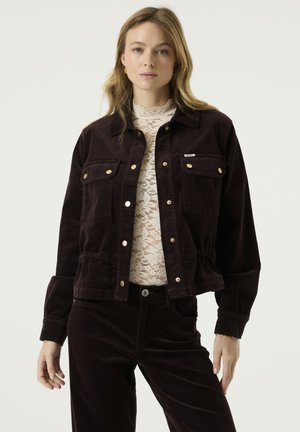 Mujer de pie con chaqueta y pantalones de pana marrón oscuro, lleva debajo una blusa de encaje blanca y mira al frente en una postura neutral.