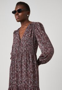 Rochie midi cu imprimeu în burgundă, având un design floral în nuanțe de albastru și alb. Dispune de mâneci lungi, decolteu în V, umeri bufanti și fustă în straturi.
