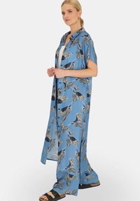 Długa niebieska koszula w stylu kimona z czarnymi wzorami liści, krótkie rękawy i boczne rozcięcia, zestawiona z białą bluzką i czarnymi sandałami ślizgaczami.