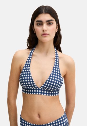 Young woman with long dark hair wearing a blue and white checkered halter bikini top against a plain light background.
