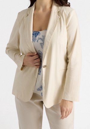 Femme portant un blazer beige par-dessus un haut blanc avec un imprimé floral bleu et un pantalon beige assorti, debout sur fond blanc.