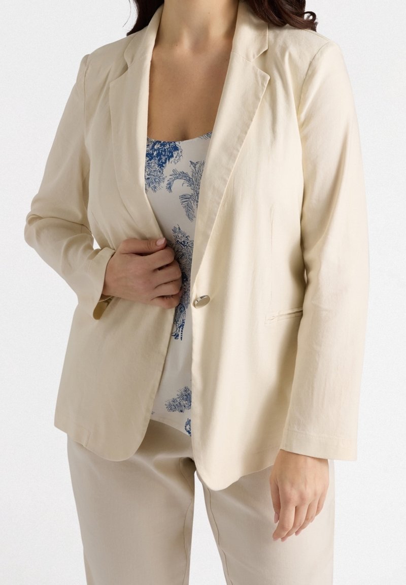 Femme portant un blazer beige par-dessus un haut blanc avec un imprimé floral bleu et un pantalon beige assorti, debout sur fond blanc.