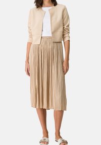 Giacca corta beige sopra una maglia bianca, abbinata a una gonna midi plissettata beige. L'outfit presenta texture lisce e dettagli di design semplice.