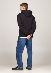 Sudadera negra con capucha de corte relajado, combinada con pantalones vaqueros azules de mezclilla que cuentan con bolsillos tipo cargo. El modelo está de pie con las manos detrás de la espalda.