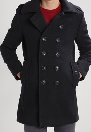 Man wearing a black double-breasted pea coat with six buttons, side pockets, and a red crew neck shirt underneath.