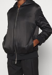 Zwarte bomberjack van satijn met een ritssluiting aan de voorkant, elastische manchetten en een contrasterende fuzzy voering in de capuchon. Zijkanten met zakken, relaxte pasvorm.