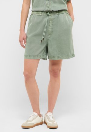 Person trägt hellgrüne Kordelzug-Shorts und weiße Sneakers, steht vor einem einfarbigen Hintergrund, Hände in den Taschen.