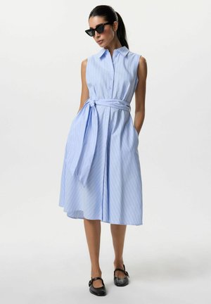 Kobieta ubrana w jasnoniebieską, paskowaną sukienkę midi bez rękawów z wiązanym paskiem w talii, czarne okulary przeciwsłoneczne i czarne buty Mary Jane, stojąca z rękami w kieszeniach.