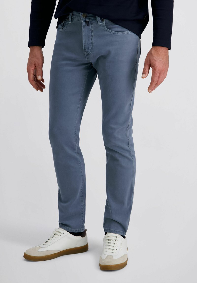 Man in slim-fit blauwe jeans en witte sneakers met bruine zolen, staande tegen een effen lichte achtergrond.
