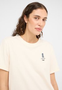 Kremowa bawełniana koszulka z krótkim rękawem, z niebieskim haftowanym wzorem dżinsów i tekstem po lewej stronie.