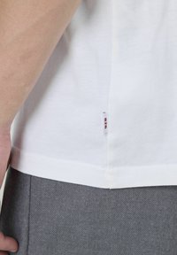 T-shirt en coton blanc à manches courtes, avec une étiquette latérale portant "BZZ". Associé à un pantalon gris texturé, mettant en valeur l'ourlet du t-shirt.