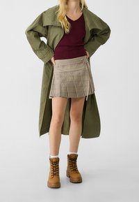 Olijfgroene lange jas, bordeauxrode aansluitende top, beige geruite mini-rok met plooien en bruine veterlaarzen met dikke zolen.