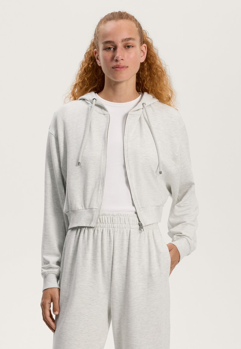 Šedá skrátená mikina na zips vyrobená z mäkkej látky s kapucňou na šnúrku, featuring hladkú textúru a bočné vrecká, nosená cez biele tričko.