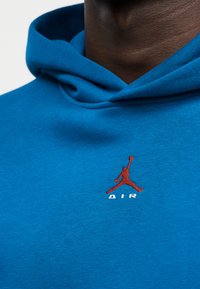 Homme portant un sweat à capuche bleu avec un petit logo rouge de joueur de basket-ball et le mot « AIR » brodé sur la poitrine.