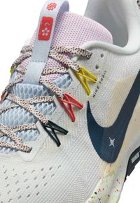 Gros plan d’une chaussure de course Nike Trail blanche avec des boucles de lacets multicolores, une semelle mouchetée et un logo floral rouge sur la languette.