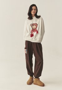 Biela mikina s potlačou medveďa s červeným šálom, hnedé nadmerné nohavice s farebným vzorovaným vreckom a béžové členkové čižmy.