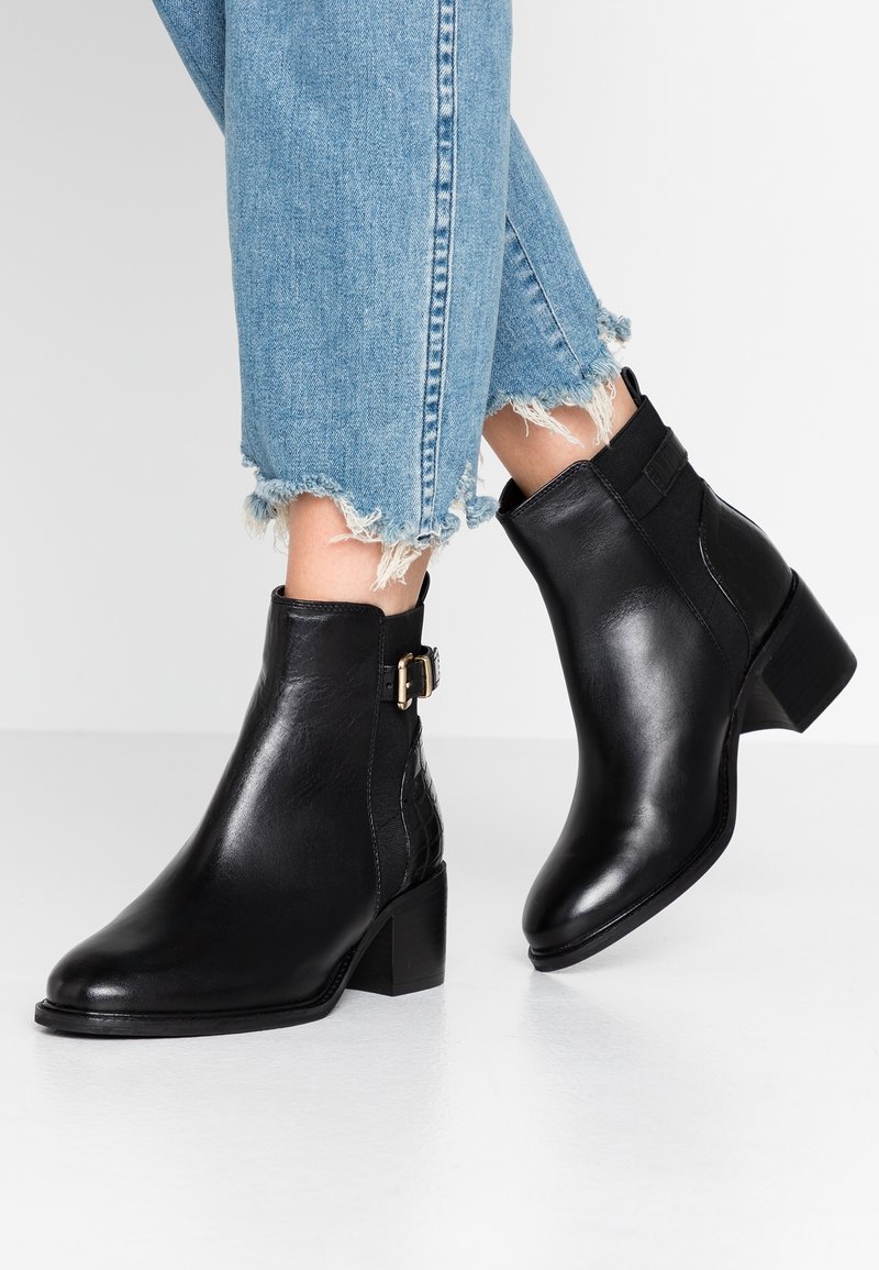 Bottines en cuir noir avec un léger talon, présentant un détail de boucle sur le côté et un panneau élastique, associées à un denim bleu clair effiloché.