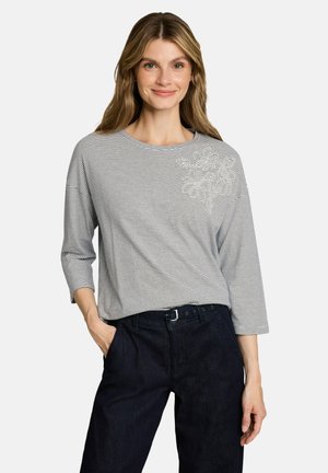 Vrouw draagt een grijs-wit gestreepte top met lange mouwen met een subtiel bloemenpatroon op de schouder, zwarte broek en glimlacht.