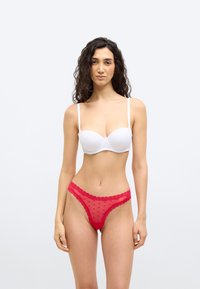 Γυναίκα με σγουρά σκουρόχρωμα μαλλιά, ντυμένη με λευκό στράπλες σουτιέν και κόκκινο δαντελένιο εσώρουχο με σχέδιο καρδιάς, στέκεται μπροστά σε απλό φωτεινό φόντο.