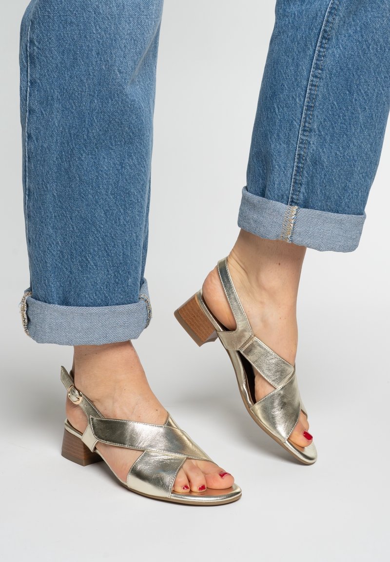 Pieds chaussés de sandales ouvertes métalliques dorées à talons blocs, associées à un jean denim bleu retroussé sur un fond uni.