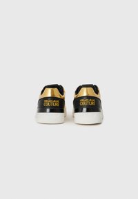 Sorte og guld sneakers med en glat læderoverflade, der har en tykkere hvid sål og "VERSACE JEANS COUTURE" trykt i gult.