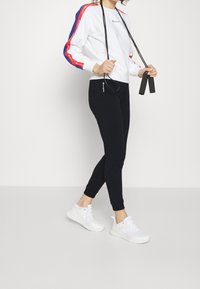 Vit sweatshirt med röda och blå ränder, svarta leggings med logotyp och vita sneakers, håller ett hopprep med svarta handtag.