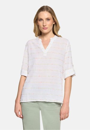 Frau trägt eine weiße, strukturierte Bluse mit hochgekrempelten Dreiviertelärmeln und hellgrünen Hosen, steht vor einem schlichten Hintergrund.