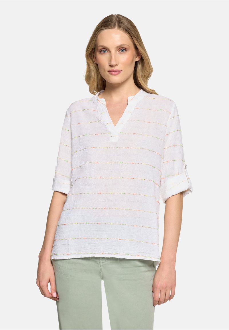 Femme portant un chemisier blanc texturé à manches trois-quarts retroussées et un pantalon vert clair, debout devant un fond uni.