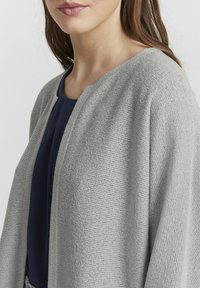 Ljusgrå stickad cardigan med öppen front, texturerad tyg och lös passform, bärs över ett mörkblått plagg. Inga knäppningar syns.