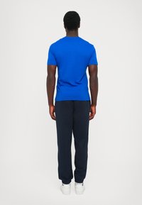 Blue cotton t-shirt with short sleeves, paired with navy sweatpants featuring a cuffed hem and white sneakers. View from the back.