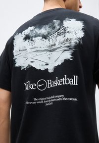 Άνδρας που φοράει μαύρο μπλουζάκι Nike Basketball με λευκό γραφικό υπαίθριου γηπέδου μπάσκετ και επωνυμη επιγραφή στην πλάτη.