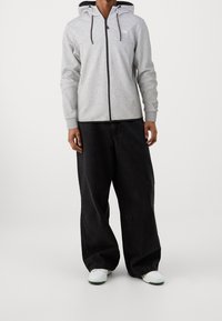 Persona che indossa una felpa grigia chiara con zip e cordini neri, pantaloni neri larghi e sneaker bianche, in piedi su uno sfondo neutro.