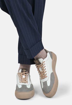 Person iført beige og grå sneakers med beige snørebånd, lysebrune ribstrikkede sokker og marineblå bukser med nålestriber og manchetter.